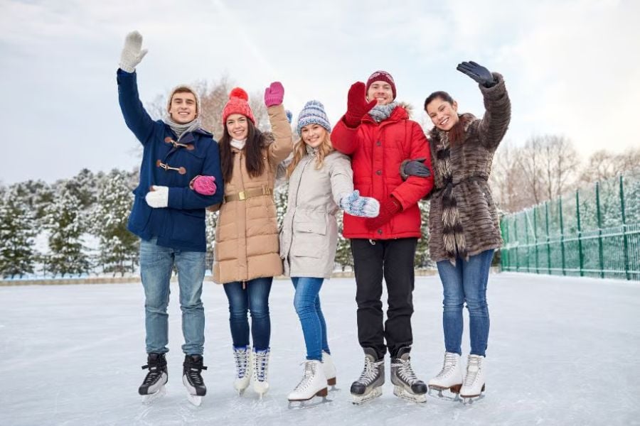 Friends wave and smile while ice skating together on a bright winter day.