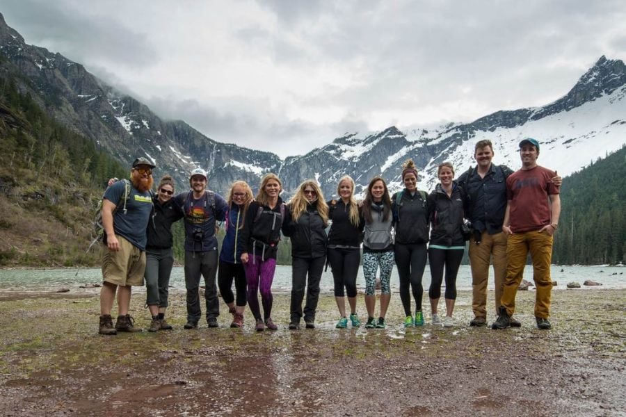 Friends pose by a mountain lake after a hike, snow-capped peaks behind them.