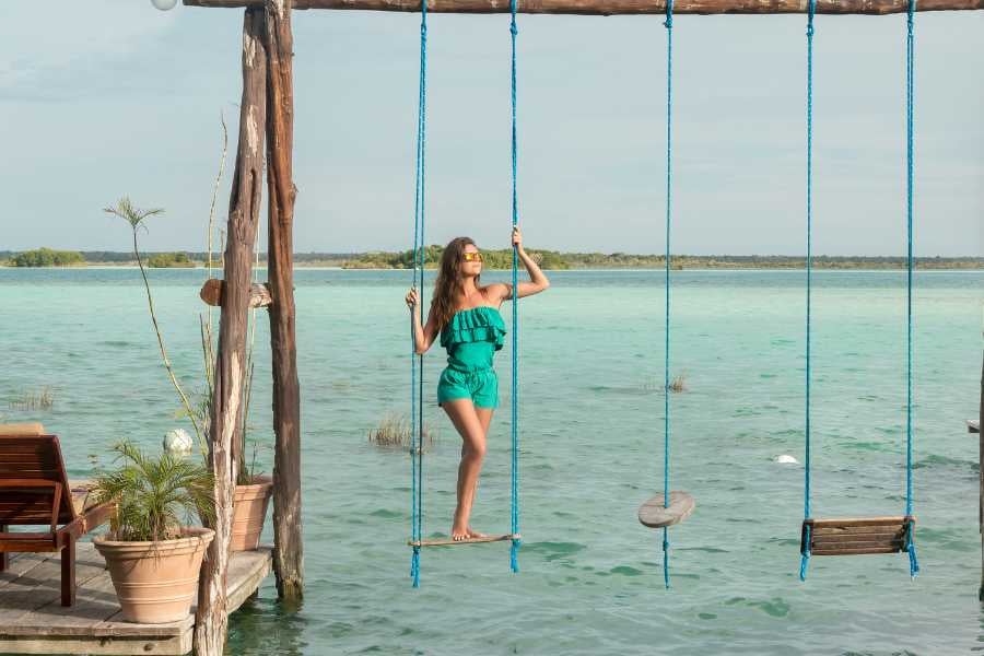 A woman poses on a swing above crystal-clear water.