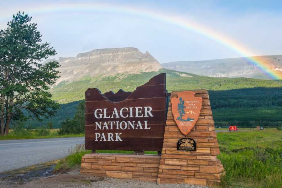 A welcoming sign at Glacier National Park, framed by mountains and a stunning rainbow.