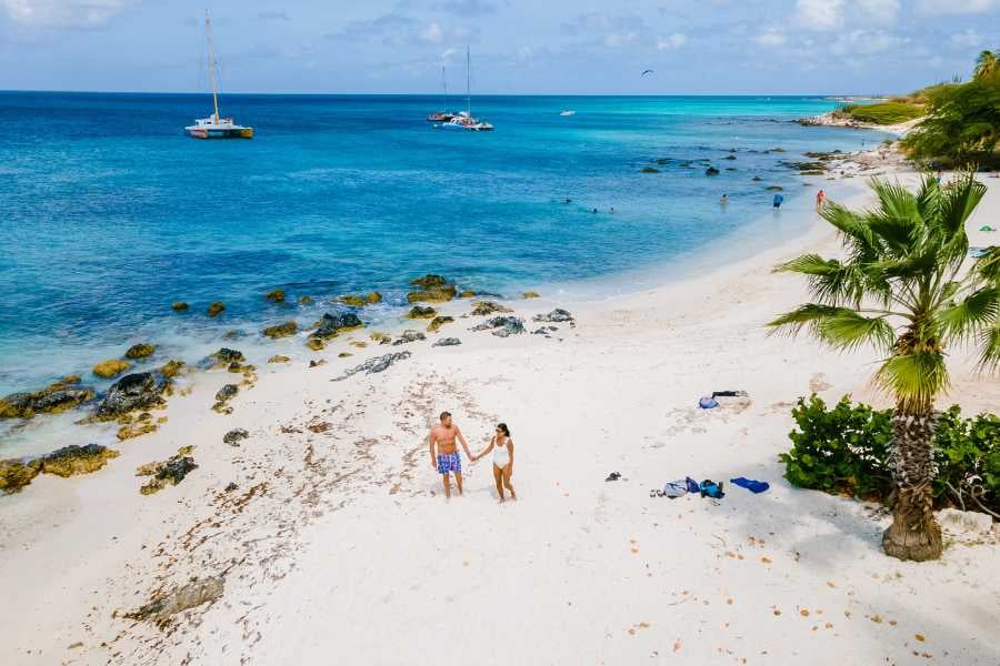 A couple walks hand in hand along a stunning white sand beach, surrounded by boats and blue waters.