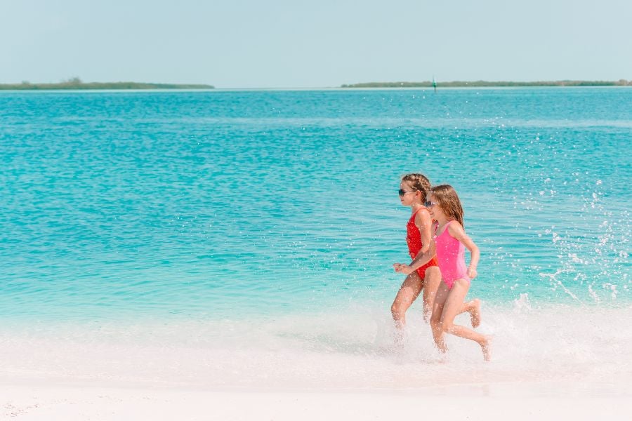 Two children run joyfully in the surf at a beautiful, tropical beach, with bright turquoise water ahead.