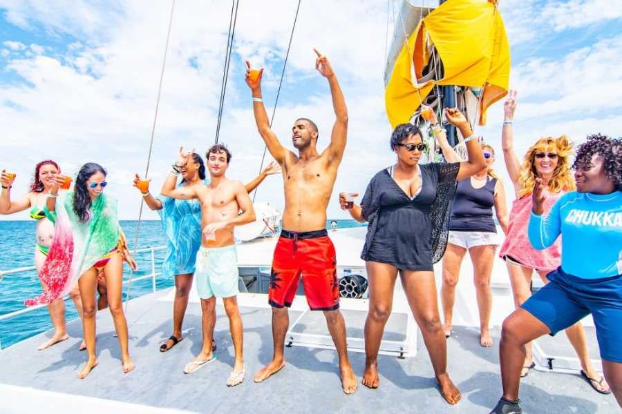 A lively group dances and celebrates on a catamaran, drinks in hand, under the sunny sky.