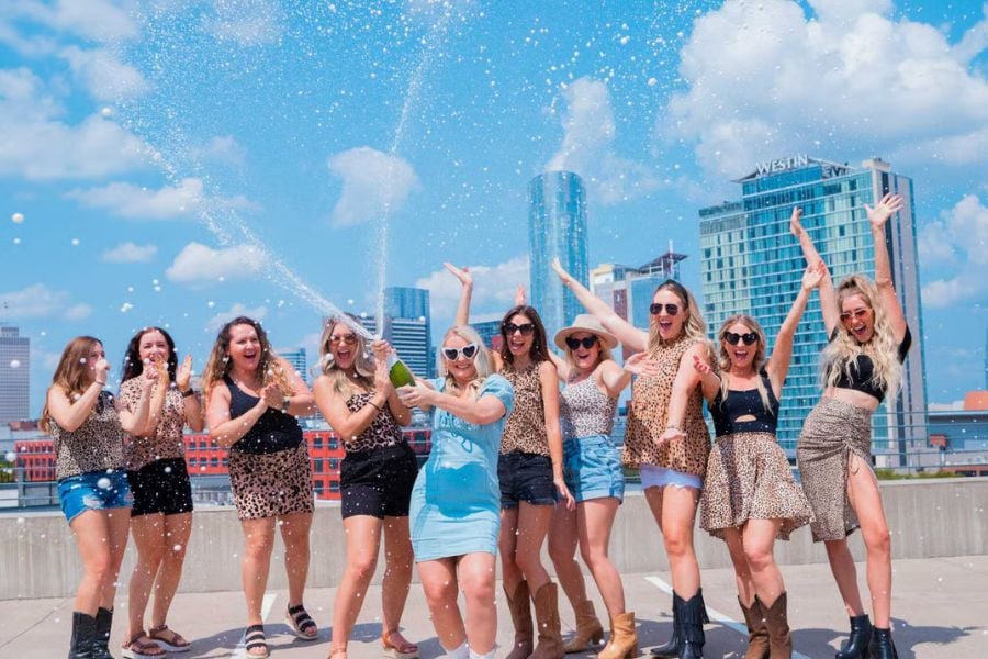Girls in leopard print outfits cheer and spray champagne on a sunny rooftop.