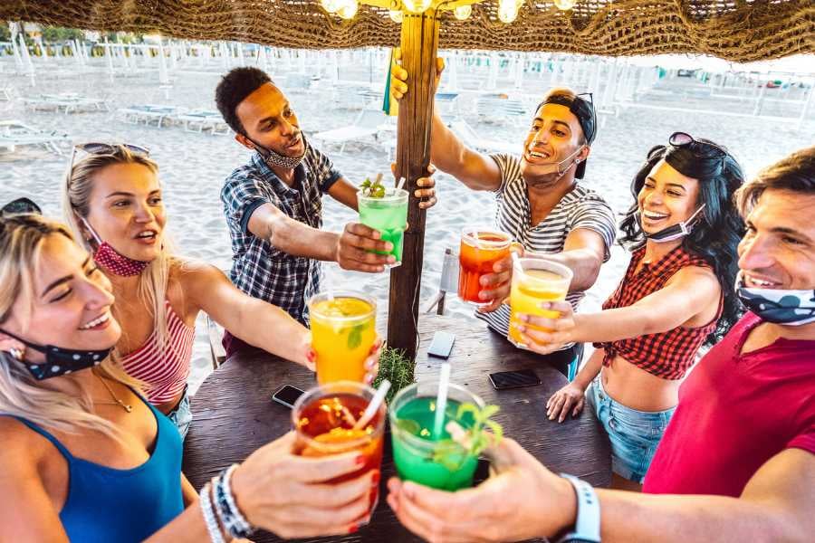 A group of friends joyfully celebrates at a beach bar, each holding vibrant, colorful drinks.