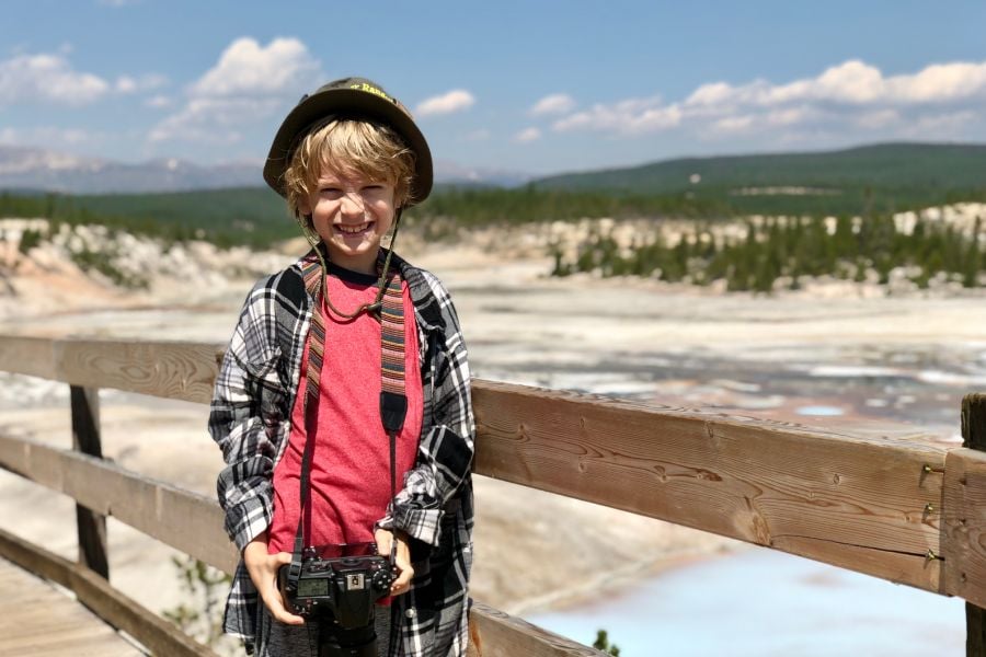A boy in a helmet smiles with a camera at Yellowstone’s boardwalk.