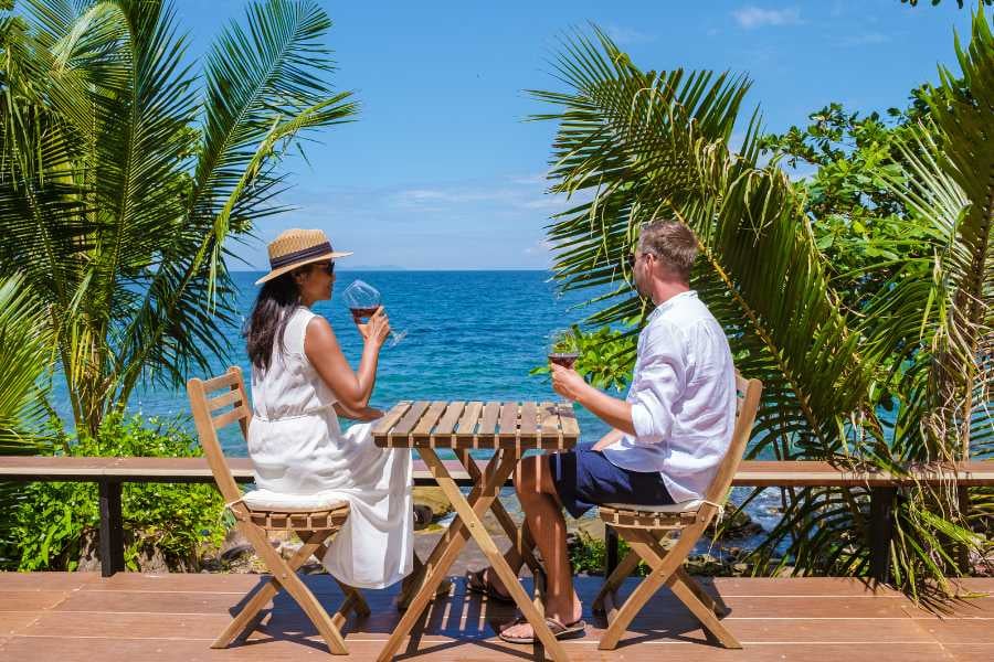 A couple enjoys a drink at a beachside table, surrounded by palm trees and ocean views.