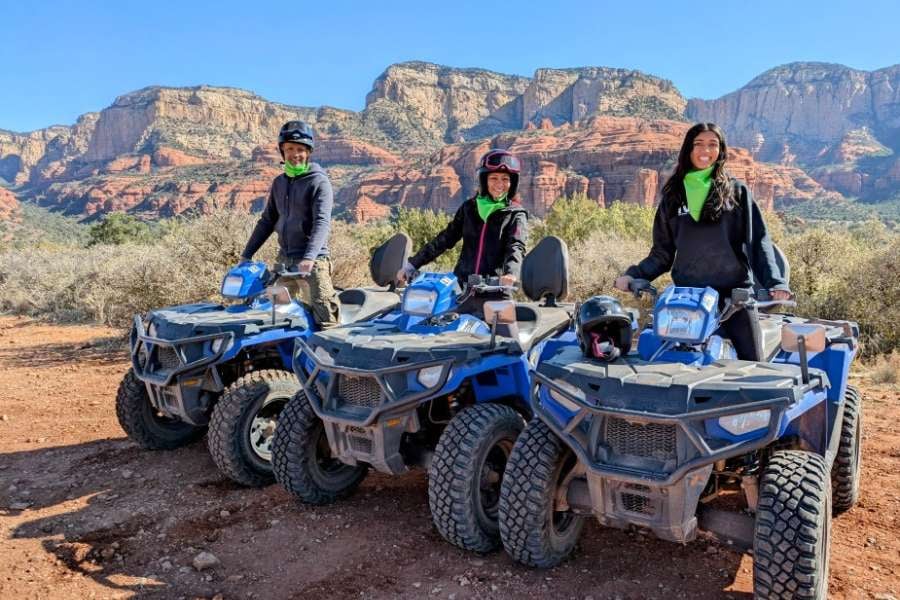 Three friends sit on their ATVs and smile in front of rocky mountains.
