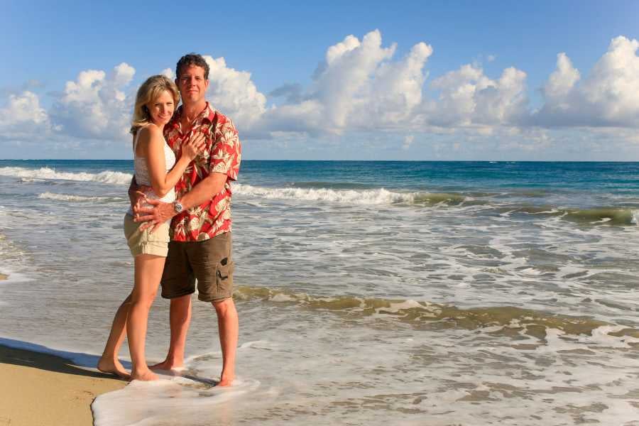 A couple stands together on a beautiful beach, smiling and embracing, with the sea in the background.