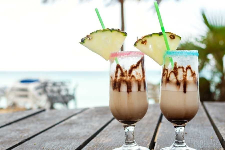 Two tropical cocktails topped with pineapple slices, sitting on a wooden table with a beach view in the background.