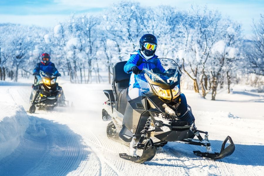 Two snowmobiles race along a sunny Breckenridge trail lined with snowy trees.