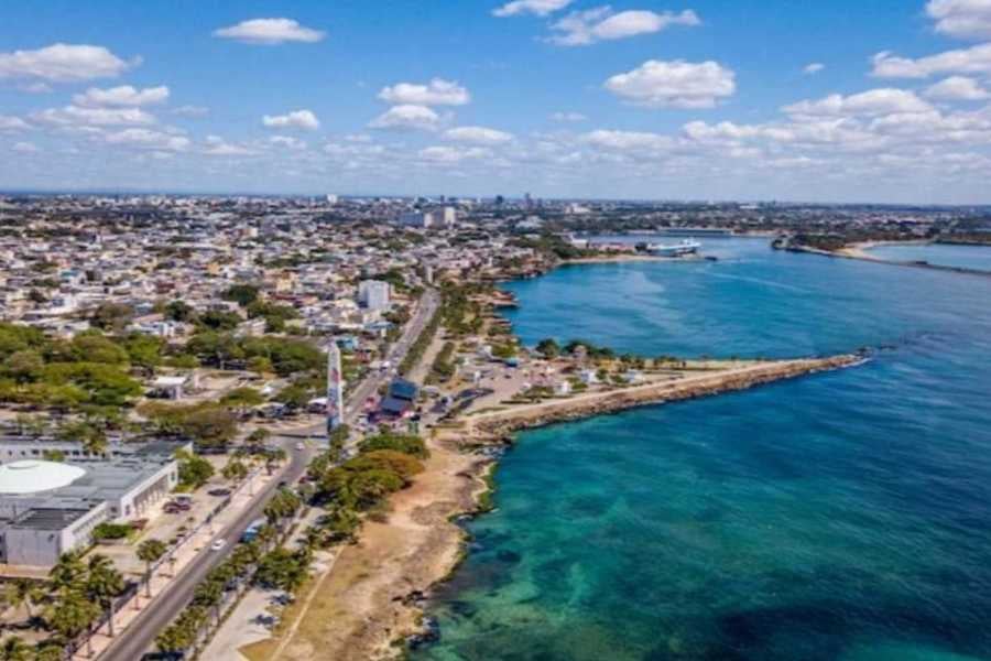 A panoramic view of a coastal city with blue water, green spaces, and a bustling road.