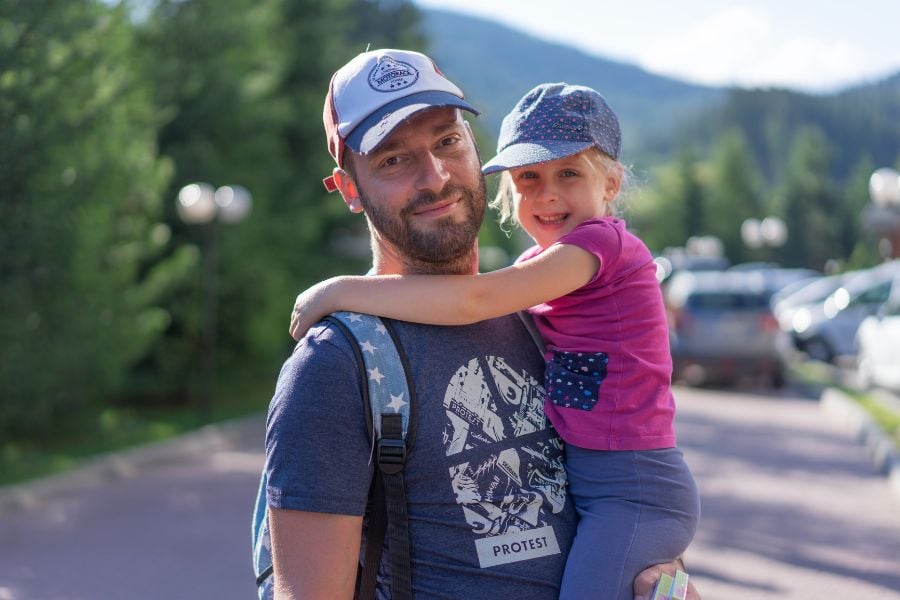 A dad smiles while carrying his daughter in a sunny mountain town.