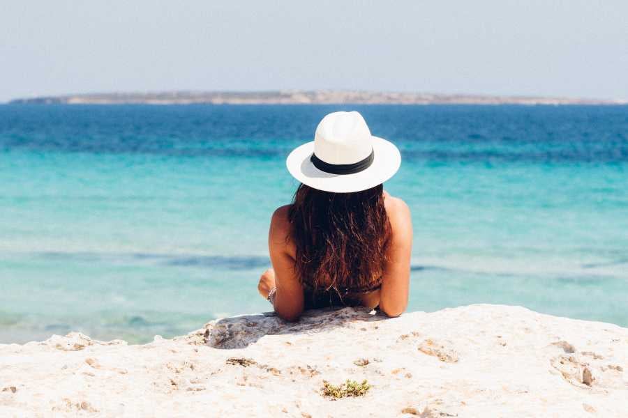 A serene moment by the turquoise sea as a woman relaxes on rocks, shaded by her stylish white hat.