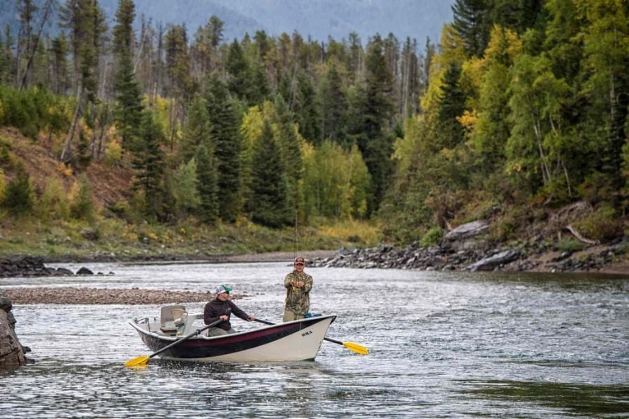Two people fishing from a drift boat in a quiet forested river setting.