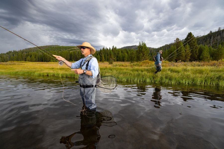 Fly fisherman stands in calm river, casting beneath stormy Yellowstone skies.