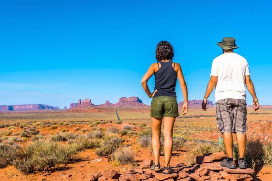 Two hikers admire the wide desert view and giant rock formations in the distance.