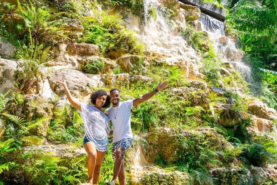 A happy couple poses in front of a breathtaking waterfall, surrounded by lush, green plants and cascading water.