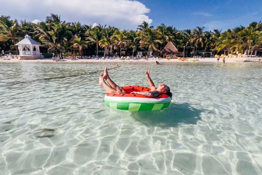 A woman relaxing on a watermelon-shaped float in clear, shallow waters surrounded by lush, tropical greenery.