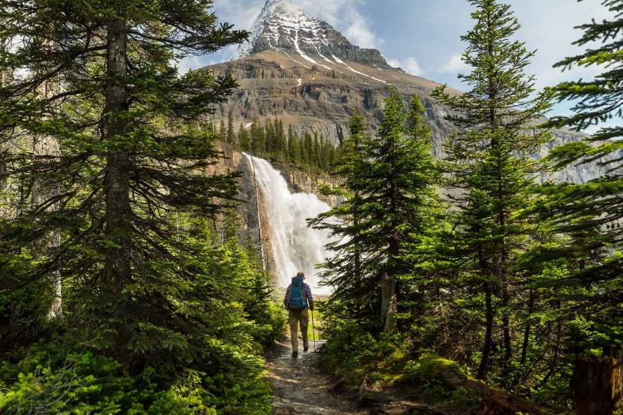 A hiker approaches a majestic waterfall beneath a towering, snow-capped mountain.