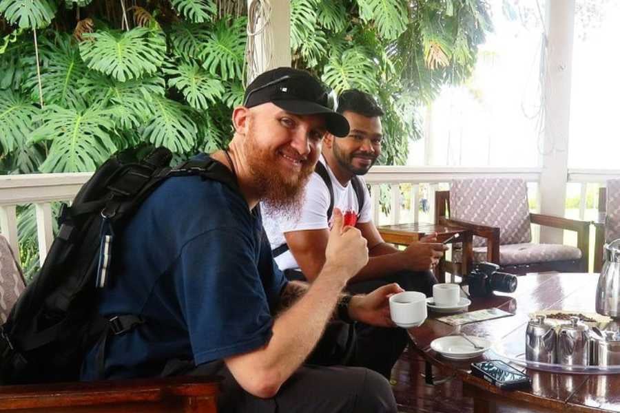 Two friends enjoy coffee on a tropical porch, smiling and relaxing with cameras ready for adventure.