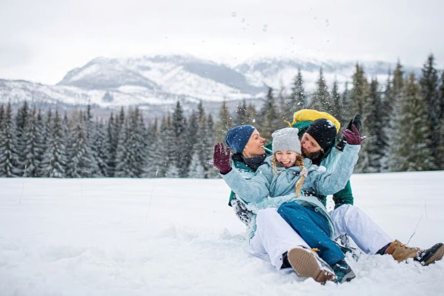 A family laughs while sliding through fresh snow in a peaceful mountain landscape.