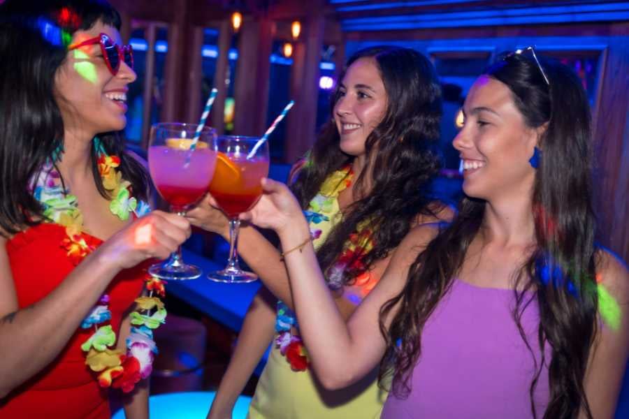 Three friends raise colorful drinks in a festive, neon-lit bar with party vibes.