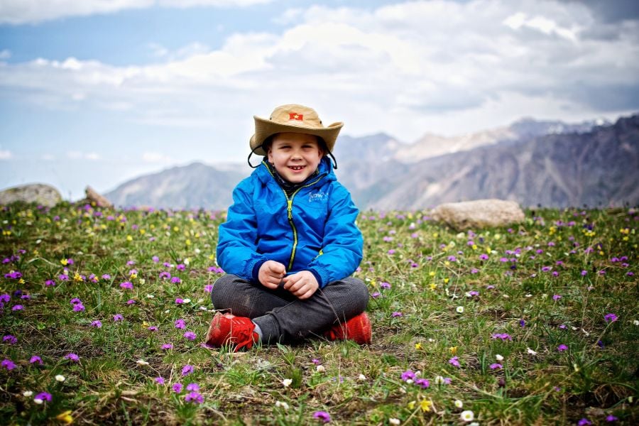 A smiling child in a blue jacket sits among mountain wildflowers.