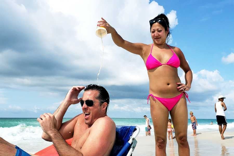 A playful beach moment—woman in a pink bikini pours a drink on a laughing man’s head.