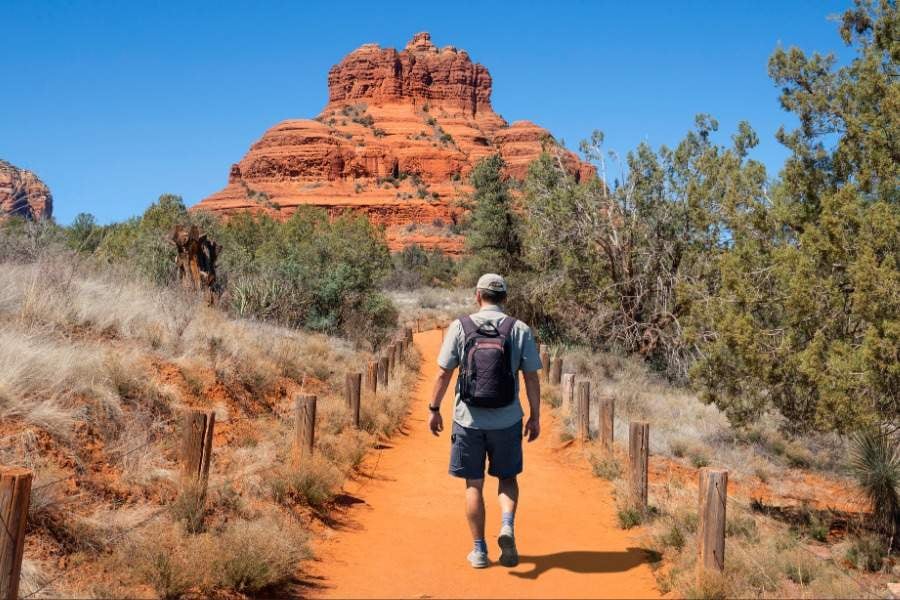  hiker walks a dusty trail toward a bright red rock formation.