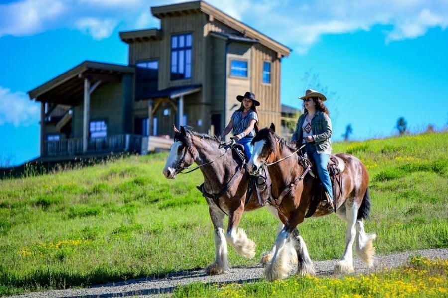 Riders on horseback pass a modern cabin in the hills.