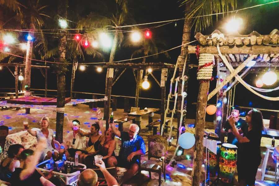 A group of friends enjoys drinks and music under festive lights at an outdoor beach bar.