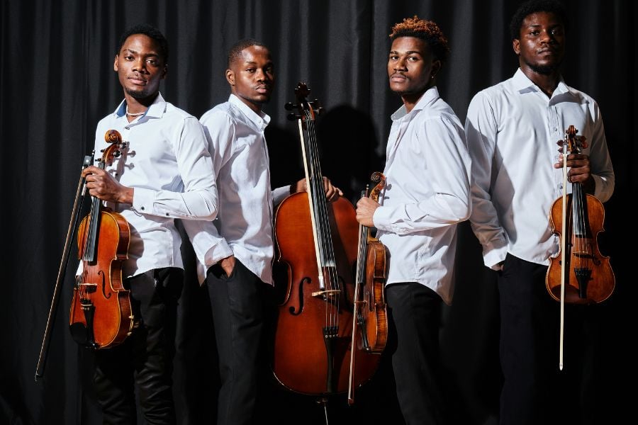 Four musicians pose with string instruments in front of a dark stage curtain.