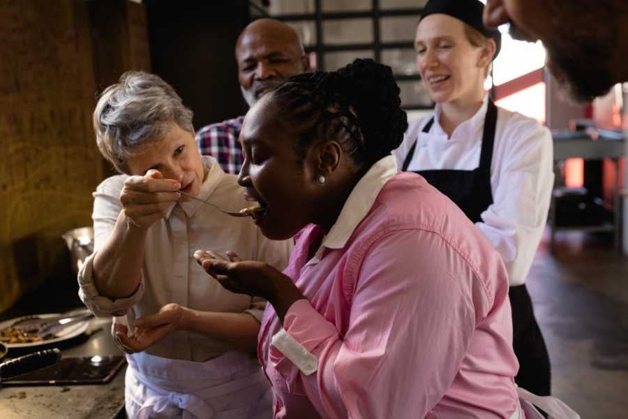 A cheerful cooking class where a woman tastes a freshly prepared dish, surrounded by smiling chefs.