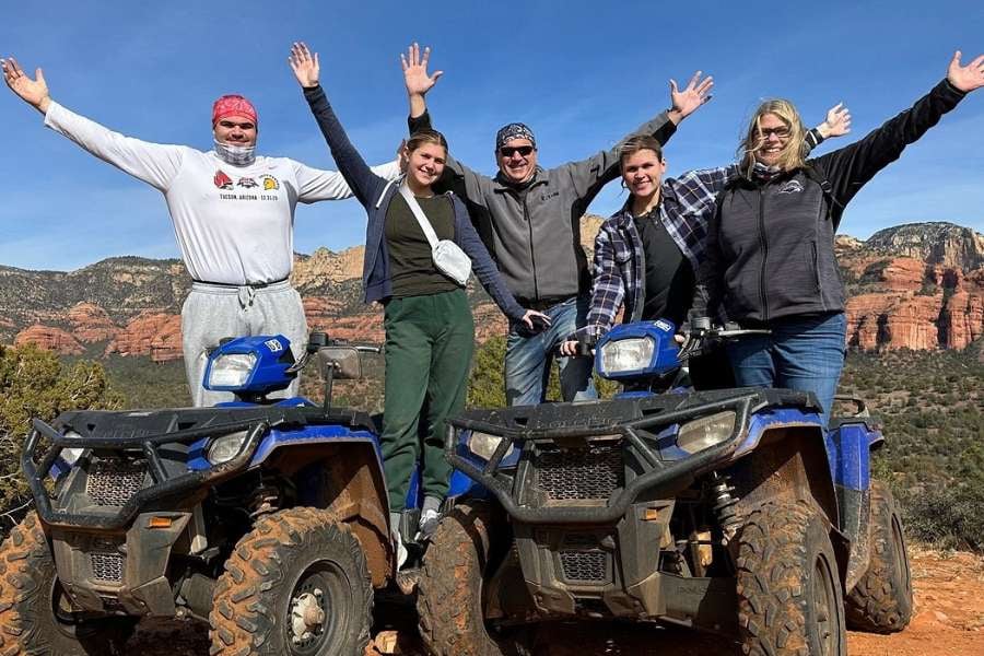 A group of six people cheers with raised arms next to two parked ATVs.