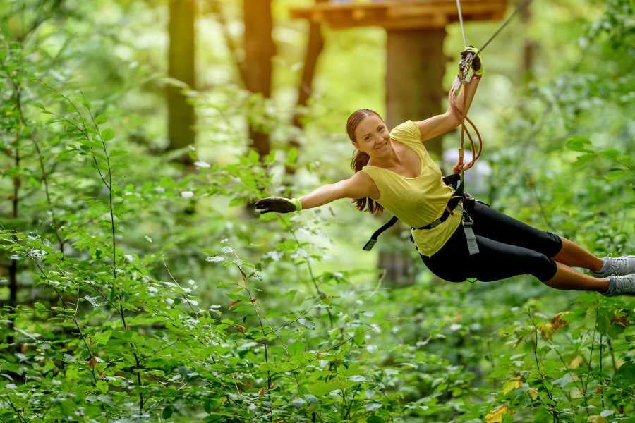 A woman glides joyfully on a zipline, surrounded by vibrant forest trees and sunlight filtering through the leaves.