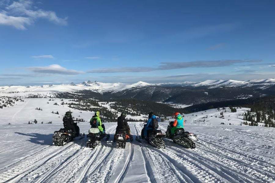 A group of snowmobilers stop on a ridge, overlooking wide snowy mountains under a clear sky.