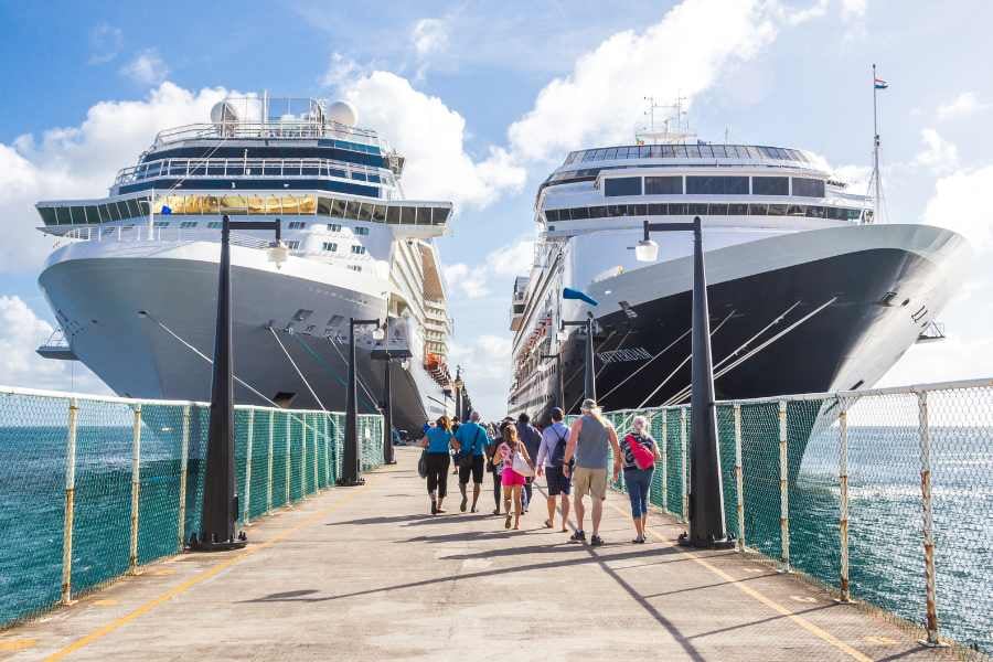 Travelers disembark from massive cruise liners docked at a bustling port under sunny skies.