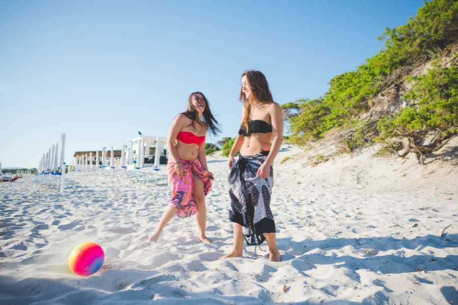 Two friends laughing and playing with a colorful beach ball on a bright, sandy beach under a sunny sky.