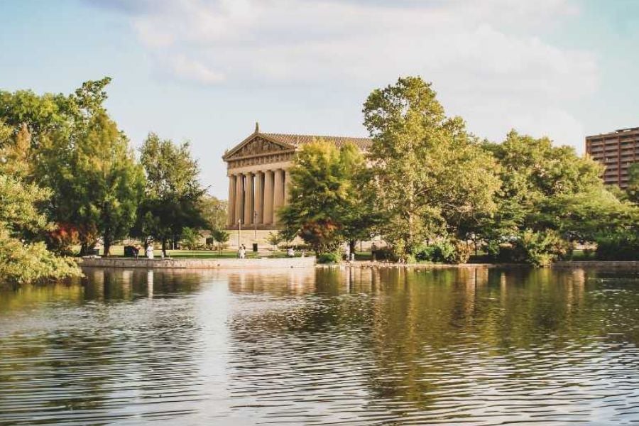 A calm lake reflects trees and a Greek-style building on a sunny day.