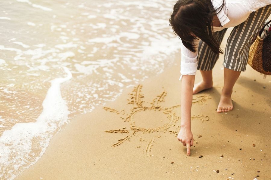 A woman draws a sun in the sand by the shoreline.