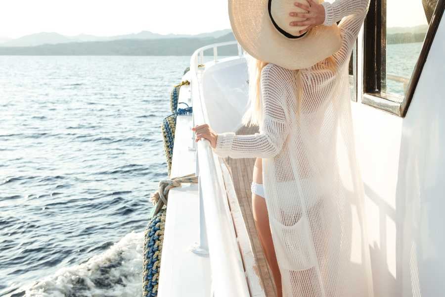 A woman in a breezy outfit gazes at the sea, holding her hat as the boat gently cruises forward.