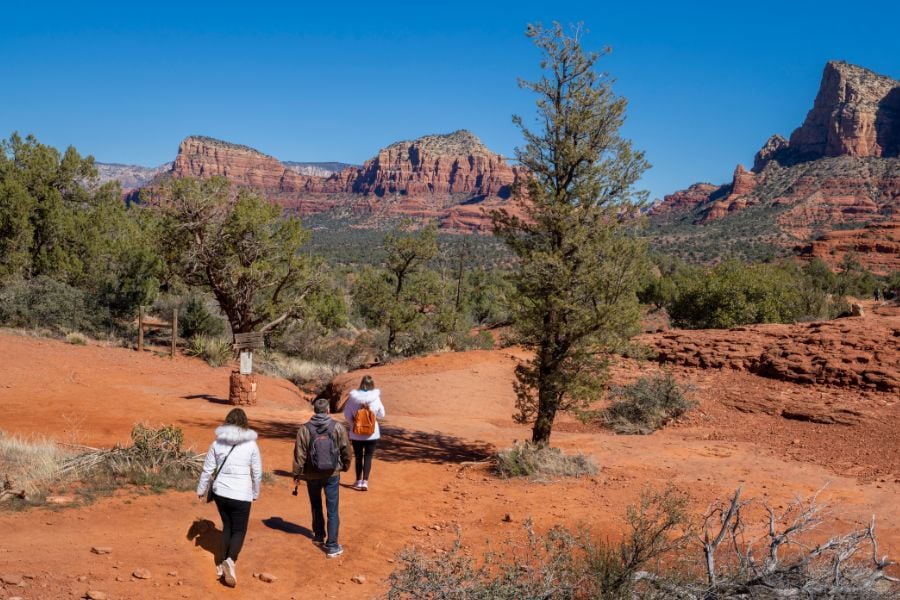 People hike a red dirt trail in Sedona with tall rock formations in the distance.