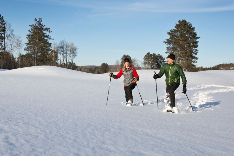Two people snowshoe across a quiet, open winter landscape, enjoying a sunny day outdoors.