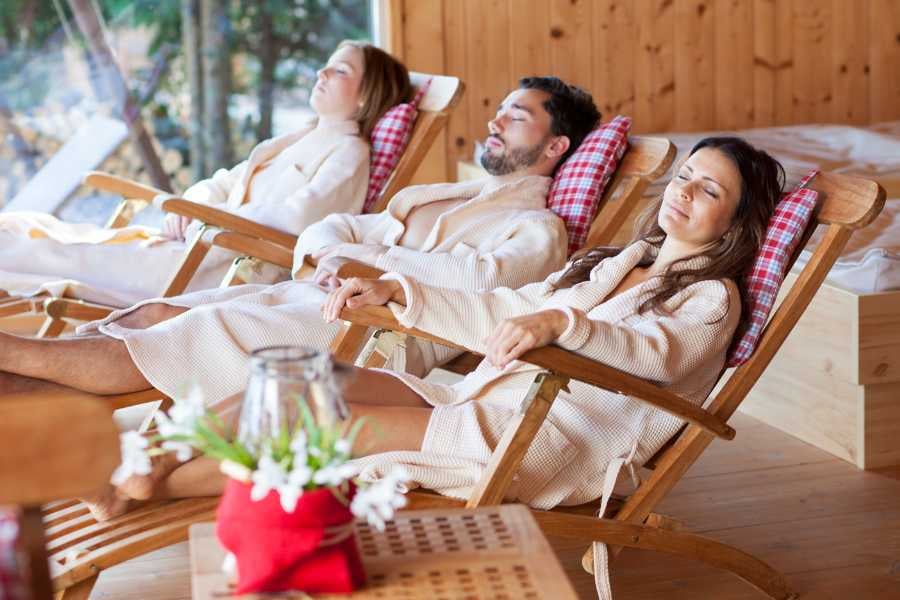 Relaxing spa scene with people reclining on wooden chairs in cozy robes, enjoying a tranquil setting.