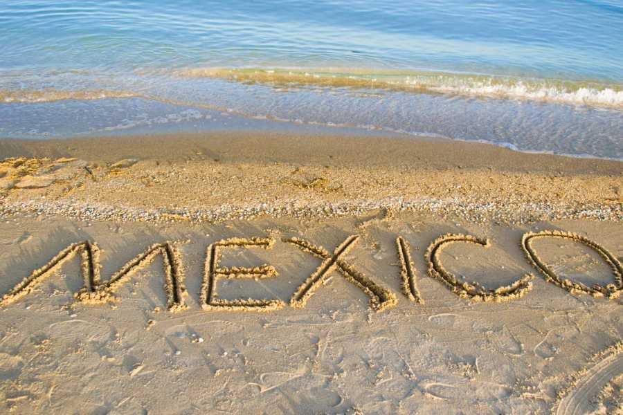 A sandy beach with “Mexico” written in the shore, as gentle waves wash up under the bright blue sky.