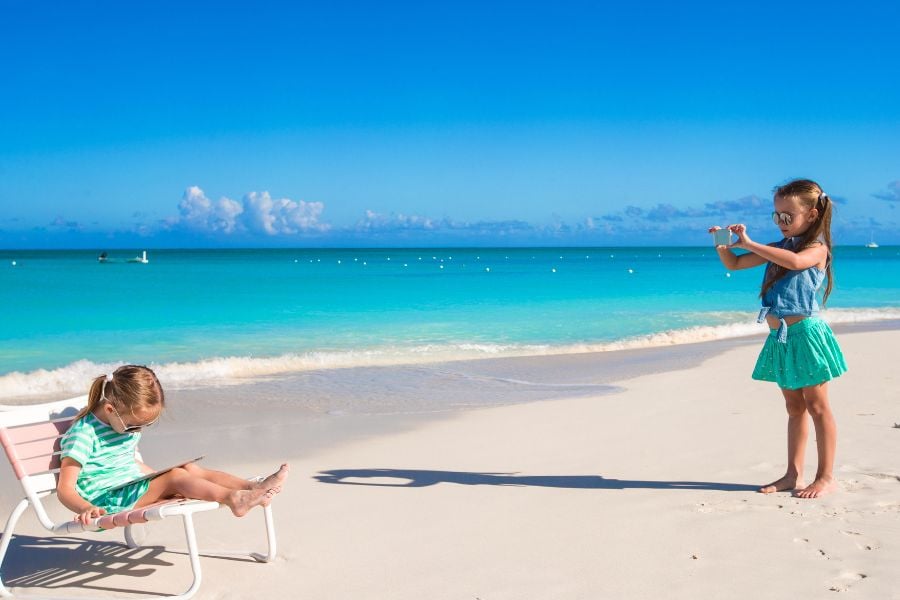 Two children enjoy a sunny beach day, one taking photos while the other relaxes in the sand.