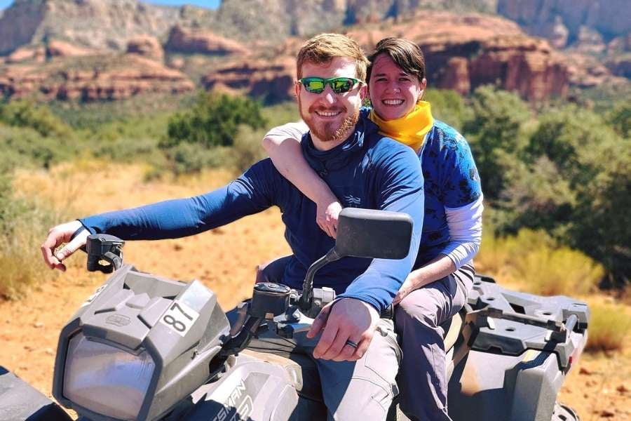 A smiling couple poses on an ATV with beautiful red rock hills behind them.