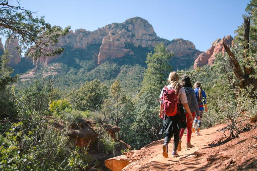 A group hikes a red dirt trail surrounded by forest and red rock cliffs.