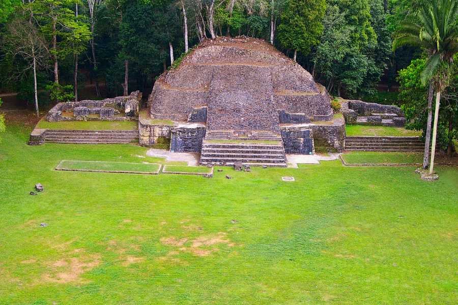 An ancient Mayan ruin surrounded by lush greenery, showcasing a stepped pyramid and open grassy space.